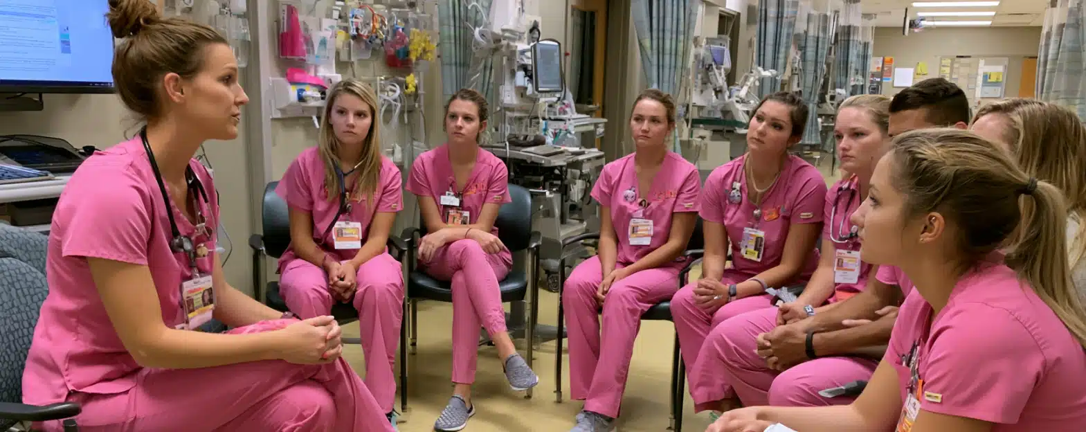 A group of nurses in pink scrubs sit in a hospital room, listening attentively to a nurse speaking.